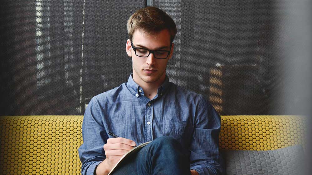 Person with glasses sitting on a couch with a textbook