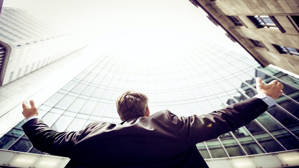 Снимка на костюмиран мъж, гледащ нагоре към стъклена сграда